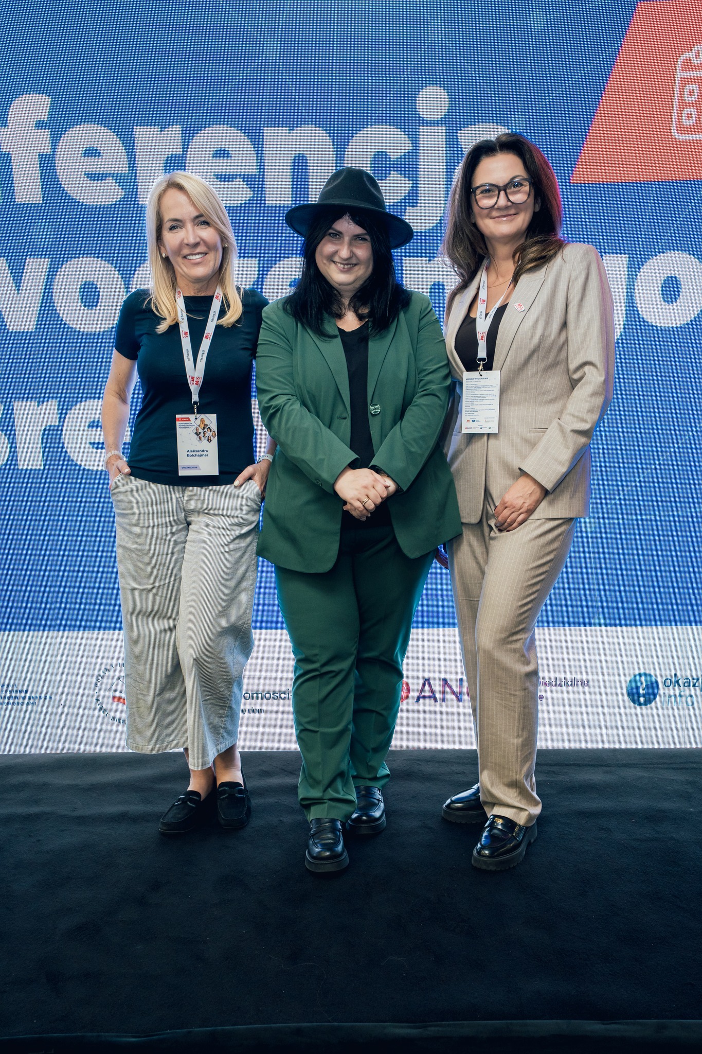 Photo of three women on stage at an MLS conference posing in front of a branded backdrop, all wearing event badges