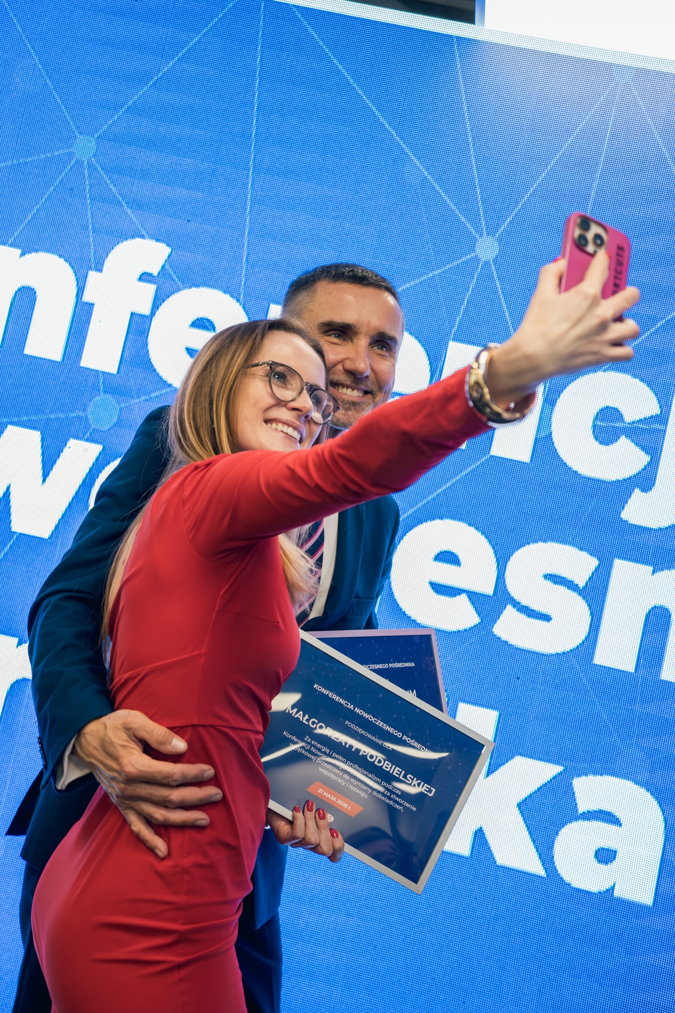 `Photo from an MLS event of a woman in a red dress taking a selfie with a man in a suit in front of a conference backdrop, holding dark blue award folders